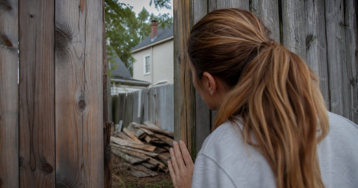 A woman peering through the gap in a wooden backyard fence toward a neighboring yard with an untidy pile of lumber, suburban houses and trees around, midday light